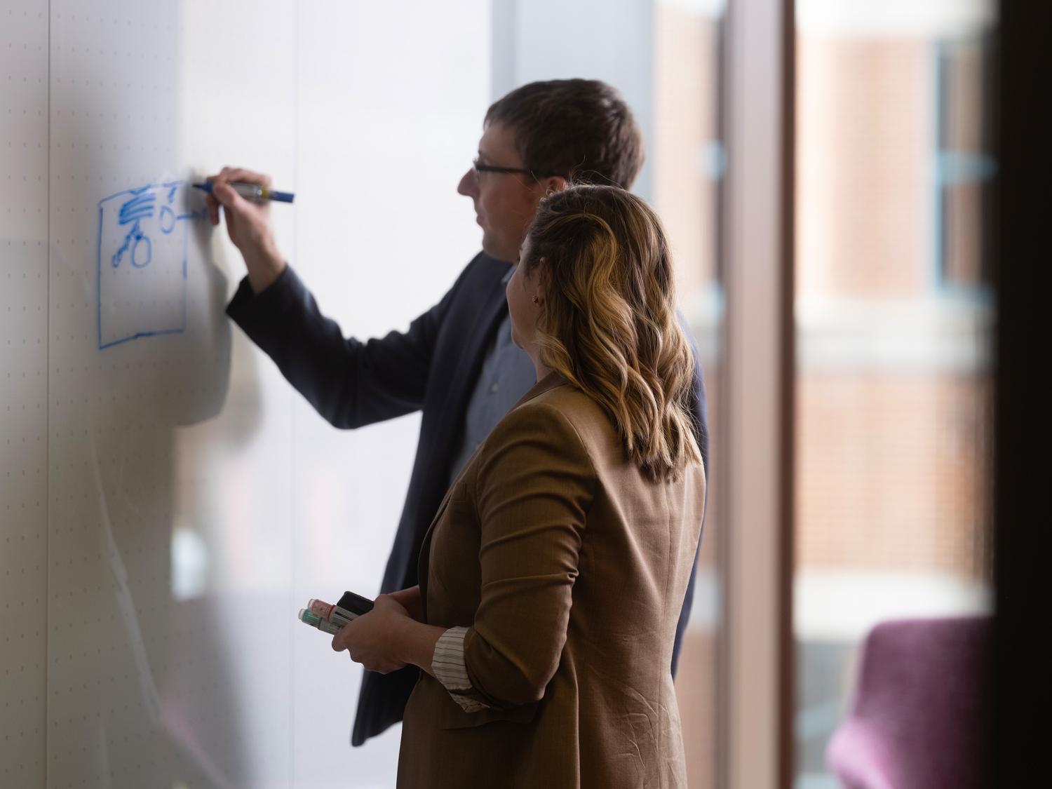 Scarlett Miller looking on as Jason Moore draws a graph on a whiteboard
